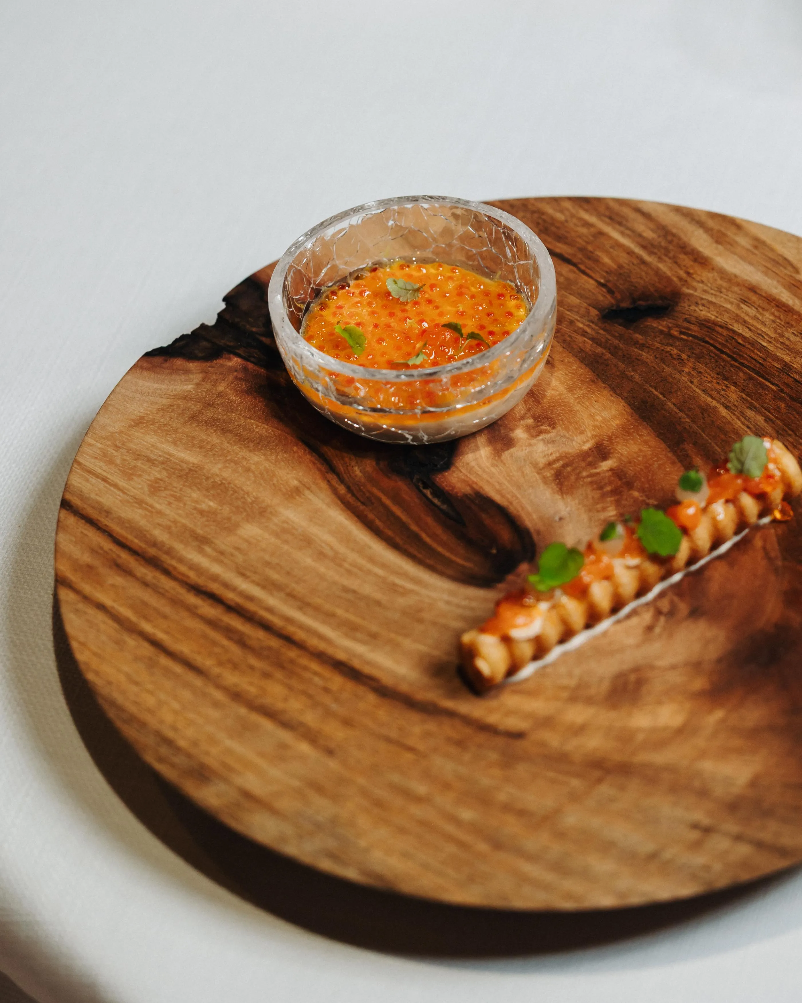 A small bowl of orange sauce with herbs and chili peppers is placed on a wooden board next to a piece of food.