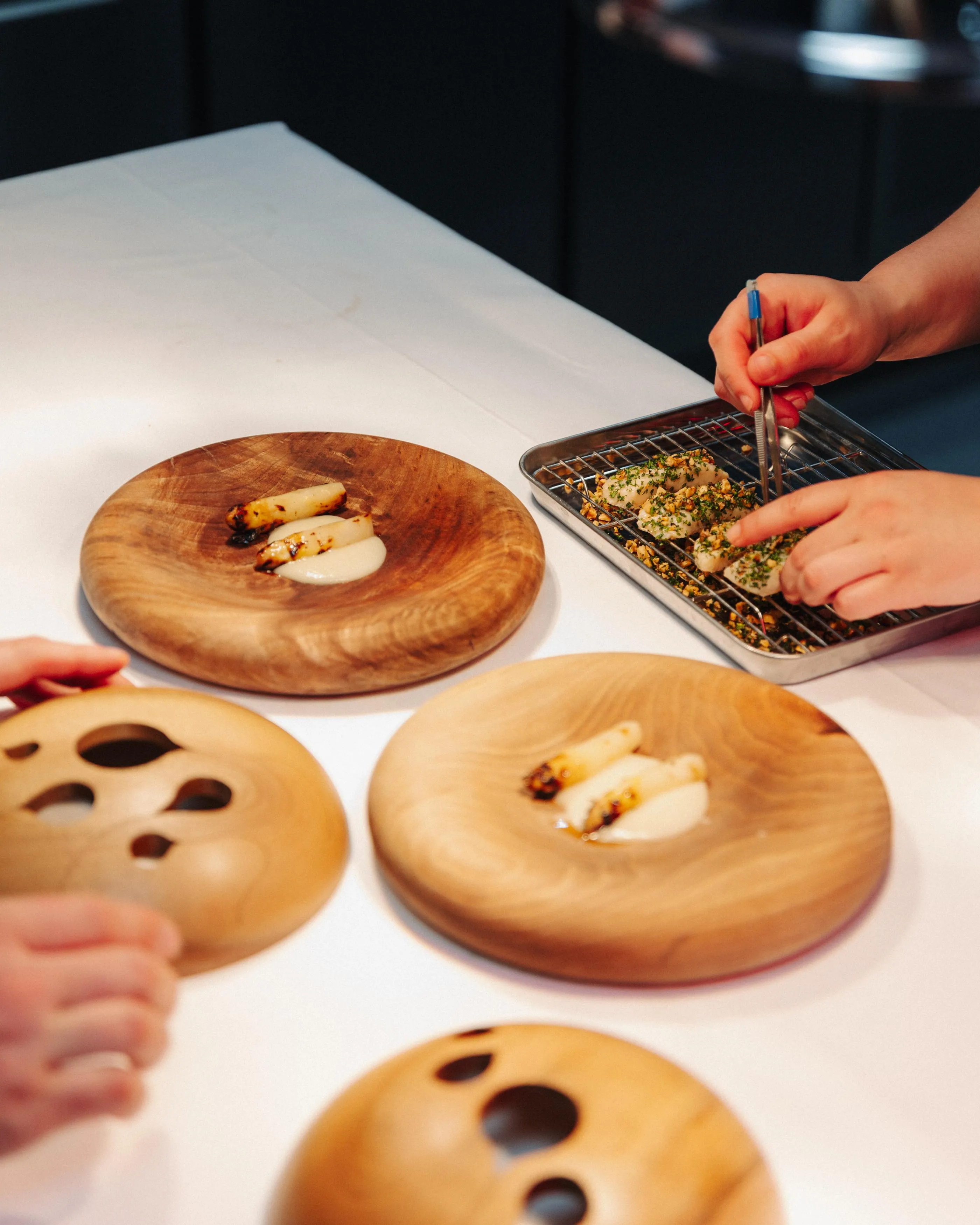 Des mains préparent des plats sur des assiettes en bois et un plateau grillagé, avec des ingrédients et des ustensiles de cuisine