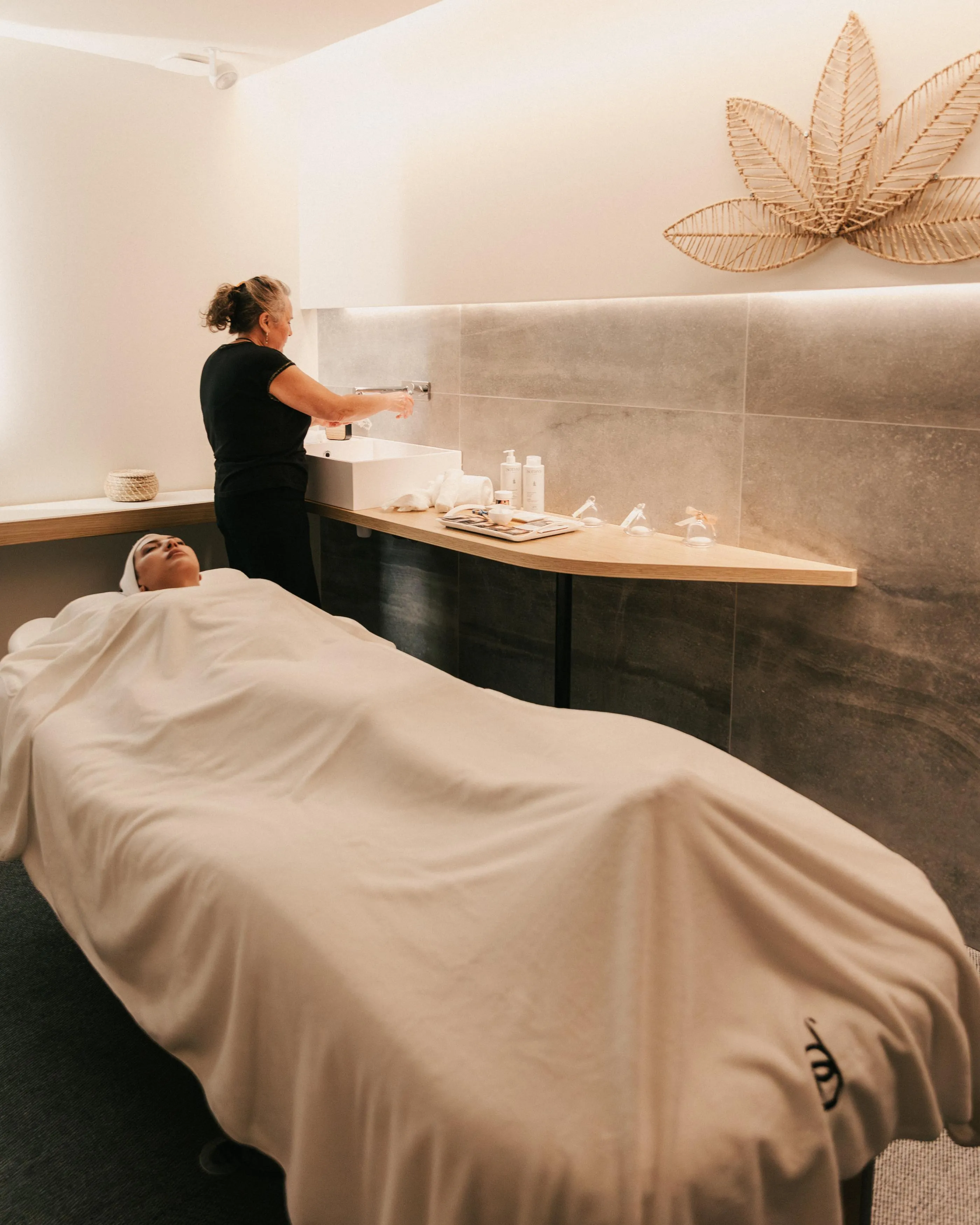 A person lying on a massage table is covered with a towel while another person stands beside them holding a bottle of product.